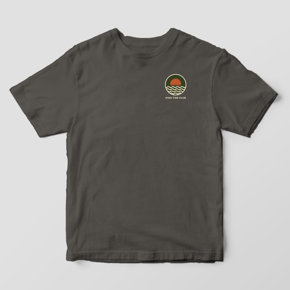 Front of a Pepper Comfort Colors 1717 tee with a small left-chest round olive badge with a rust half-sun and cream waves above HIGH TIDE CLUB.