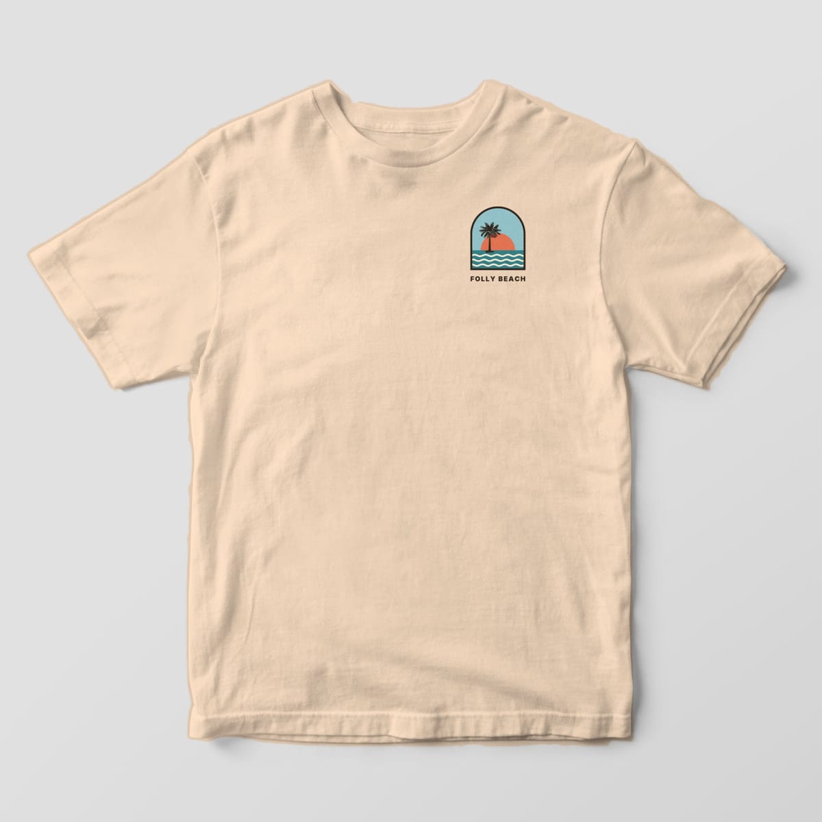 Front of an Ivory Comfort Colors 1717 tee with a small left-chest arch with a palmetto, coral sun and teal waves above the words FOLLY BEACH.