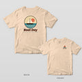Ivory Comfort Colors tee shown back and front: a round sailboat badge over Boat Day on the back and a small coral sailboat over Boat Day on the left chest.