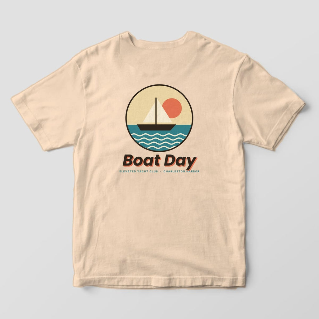 Back of an Ivory Comfort Colors 1717 tee with a round badge of a sailboat on teal water under a coral sun, above Boat Day and ELEVATED YACHT CLUB · CHARLESTON HARBOR.