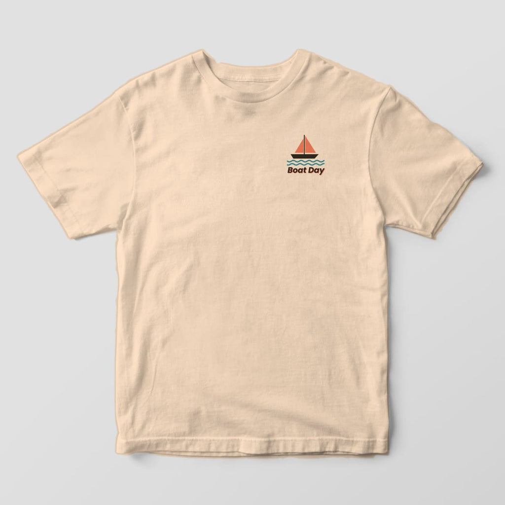 Front of an Ivory Comfort Colors 1717 tee with a small left-chest sailboat with coral sails and teal waves above the words Boat Day.
