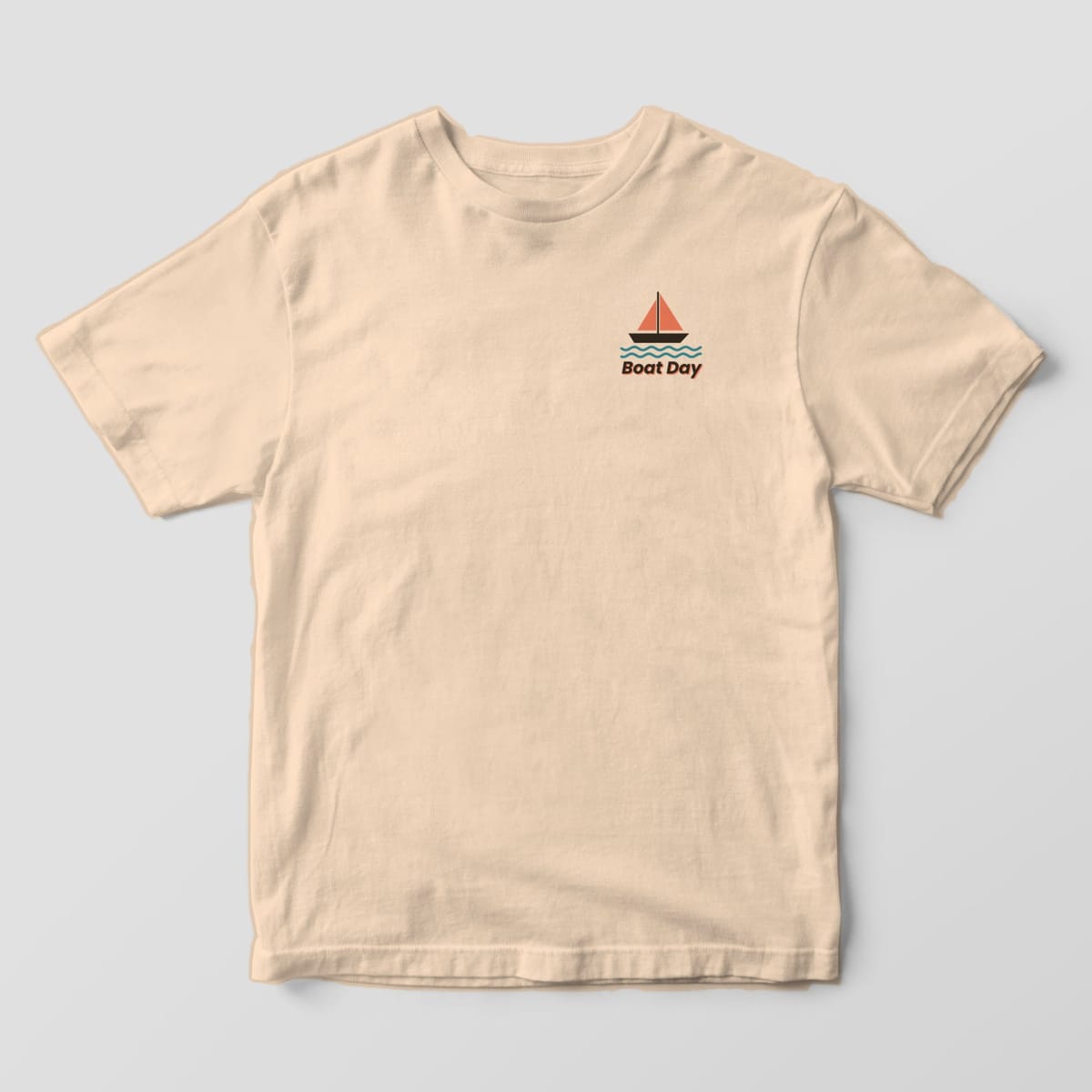 Front of an Ivory Comfort Colors 1717 tee with a small left-chest sailboat with coral sails and teal waves above the words Boat Day.