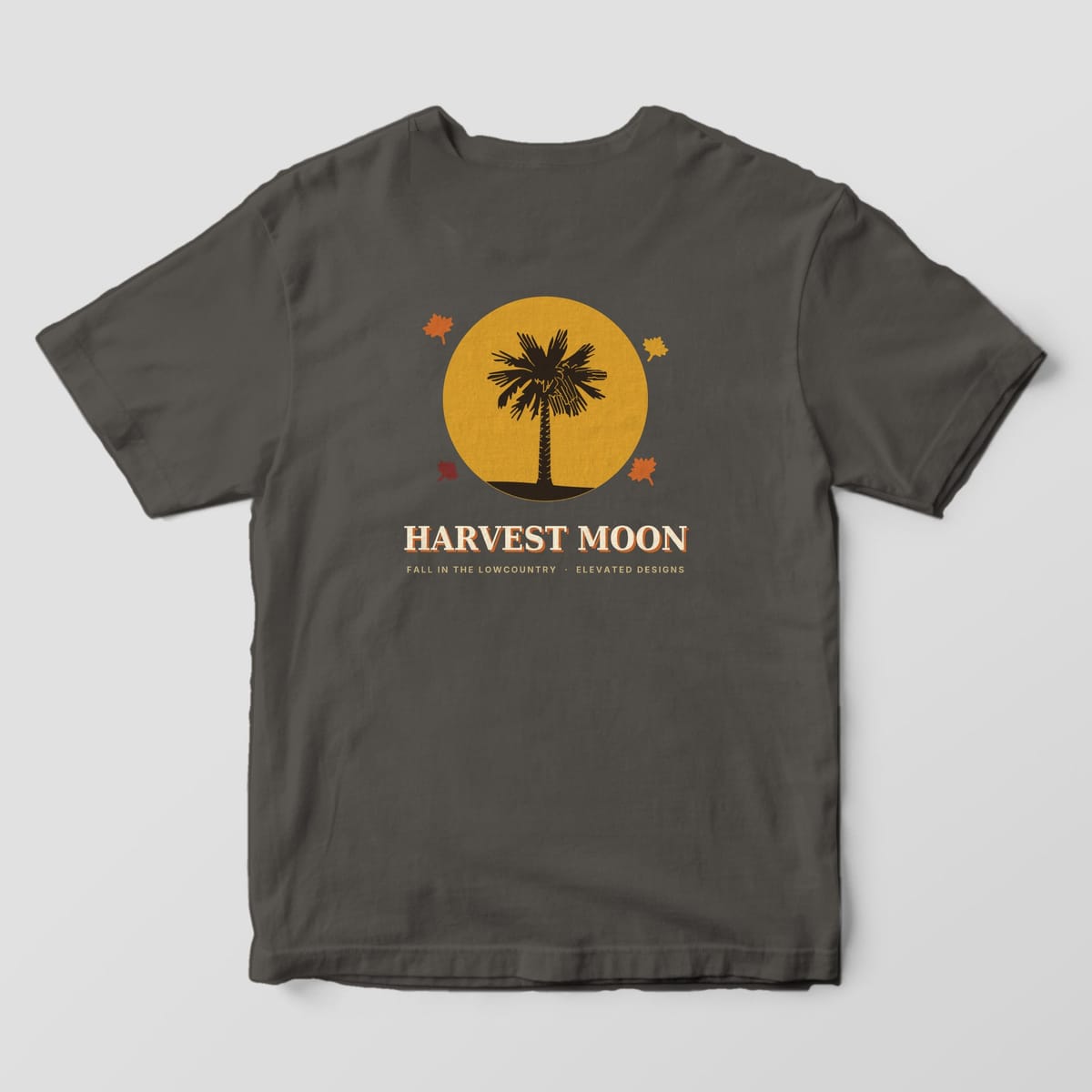 Back of a Pepper Comfort Colors 1717 tee with a dark palmetto silhouette in front of a large gold moon with four autumn leaves, above HARVEST MOON and FALL IN THE LOWCOUNTRY · ELEVATED DESIGNS.