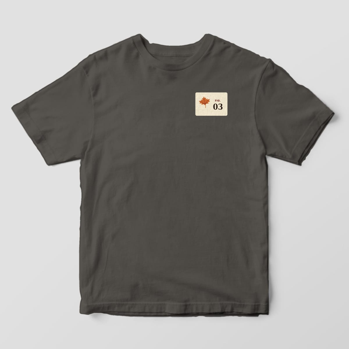 Front of a Pepper Comfort Colors 1717 tee with a small left-chest cream card with a rust maple leaf and the label FIG. 03.
