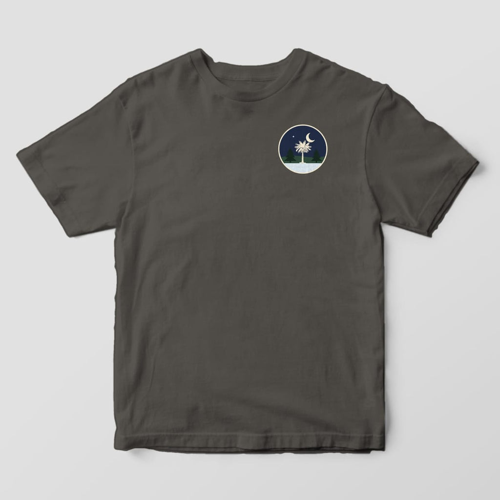 Front of a Pepper Comfort Colors 1717 tee with a small round left-chest night scene with a crescent moon, a star, a cream palmetto, two pine trees and snow.