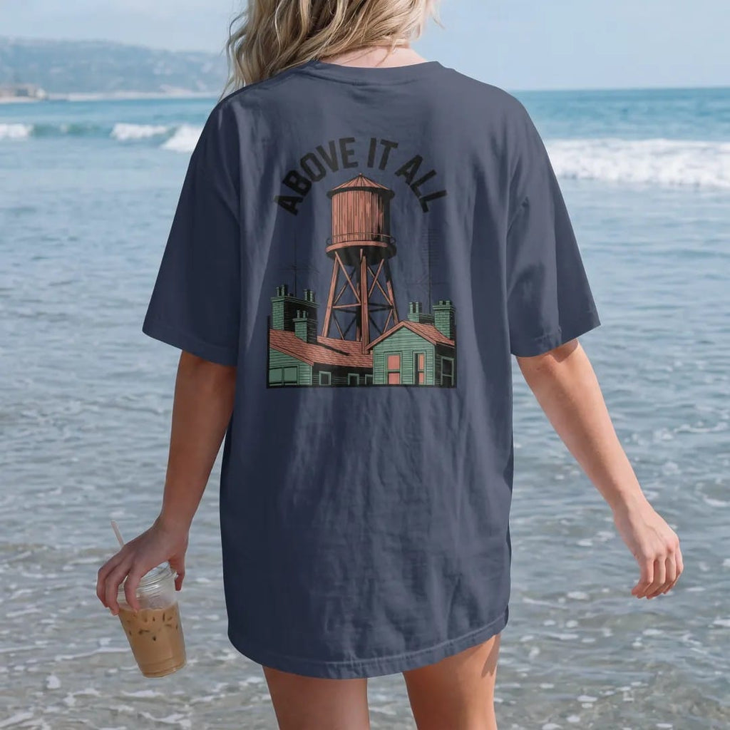 Woman on the beach in a Navy tee, back view: a wooden rooftop water tower over brownstone roofs with Above It All arched across the top in bone white, orange and green.