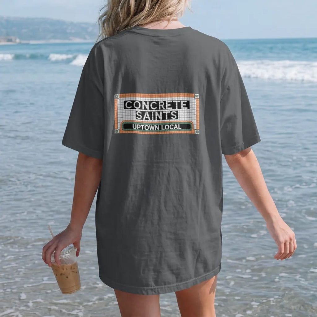 Woman on the beach in a Graphite tee, back view: a subway-style mosaic tile name tablet spelling Concrete Saints over a smaller Uptown Local band, in bone white, green, orange and yellow tiles.