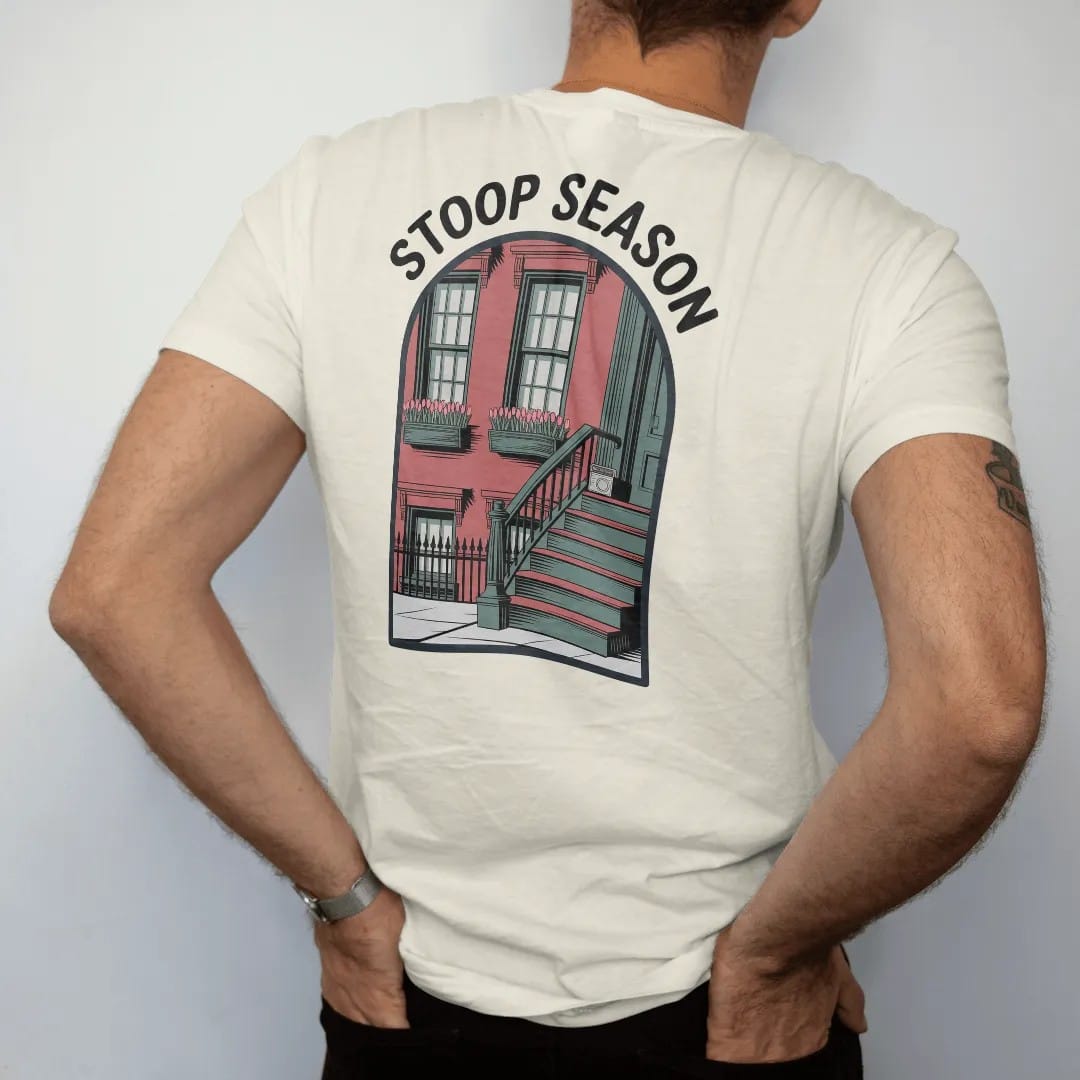 Man wearing a Ivory tee, seen from the back: a brownstone stoop with tulip window boxes and a radio on the steps under hand-painted Stoop Season lettering, in charcoal, brick red, green and navy.