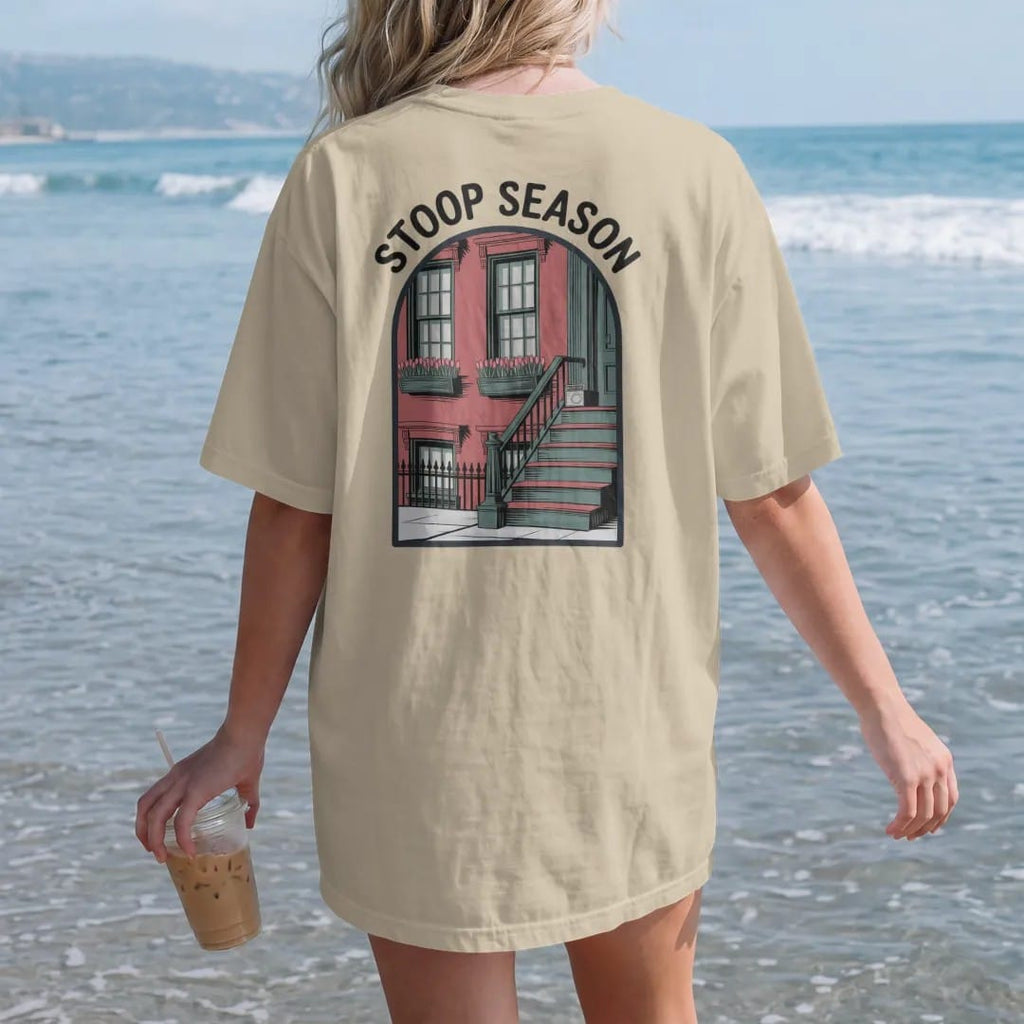 Woman on the beach in a Sandstone tee, back view: a brownstone stoop with tulip window boxes and a radio on the steps under hand-painted Stoop Season lettering, in charcoal, brick red, green and navy.