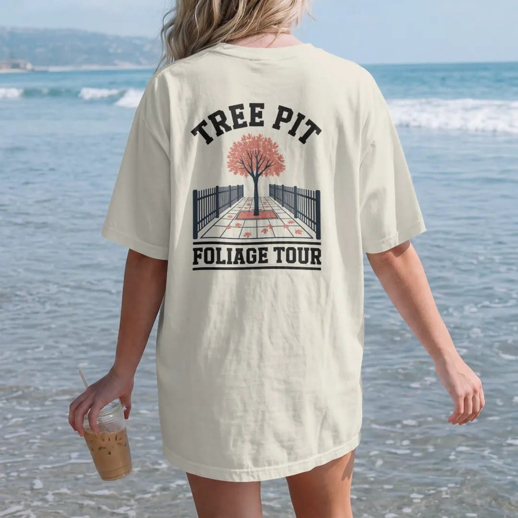 Woman on the beach in a Ivory tee, back view: a single sidewalk tree in a fenced pit dropping orange leaves, under Tree Pit and over Foliage Tour, in charcoal, brick red, green and navy.