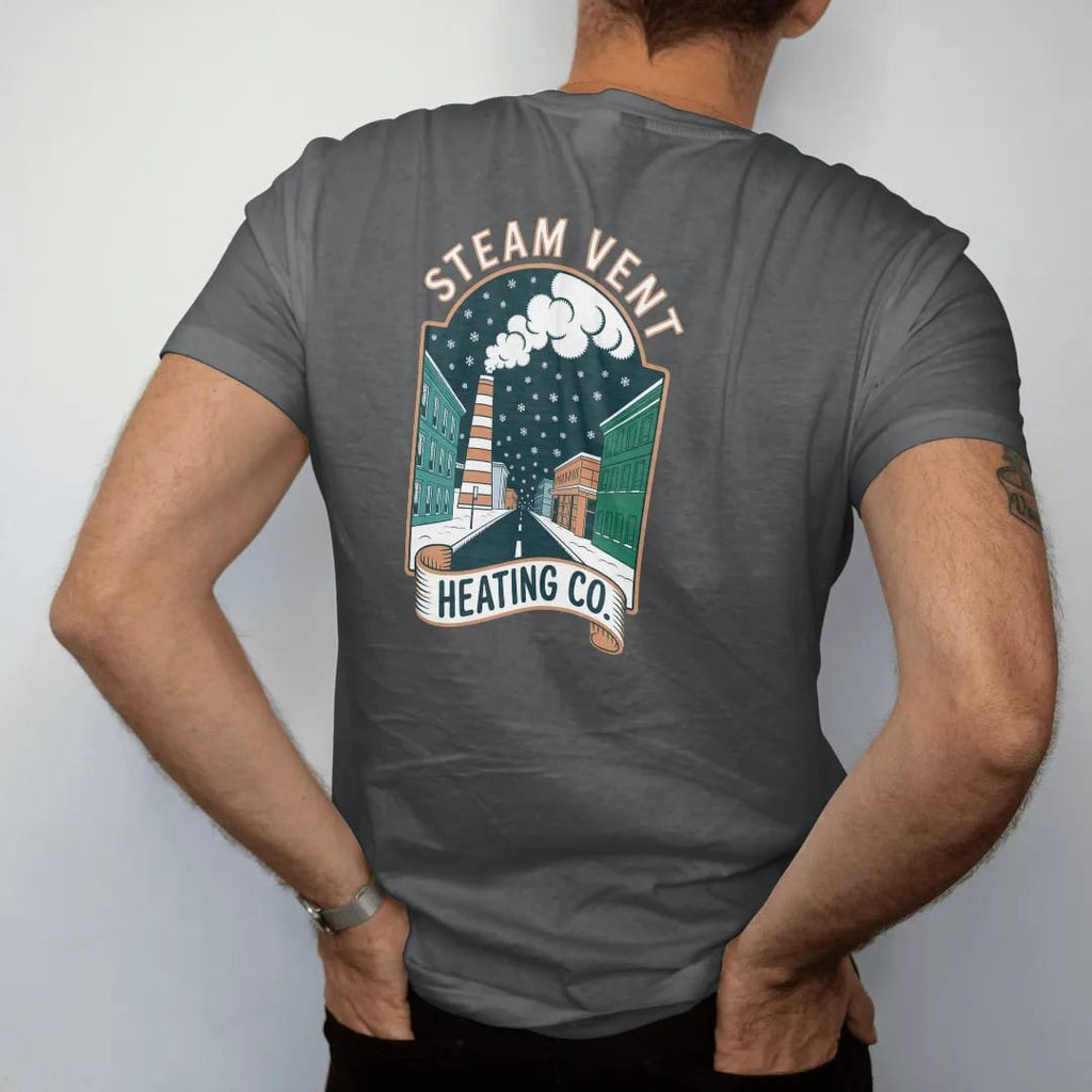 Man wearing a Graphite tee, seen from the back: a striped steam stack puffing clouds in falling snow with Steam Vent arched above a Heating Co. ribbon, in bone white, orange, yellow and green.