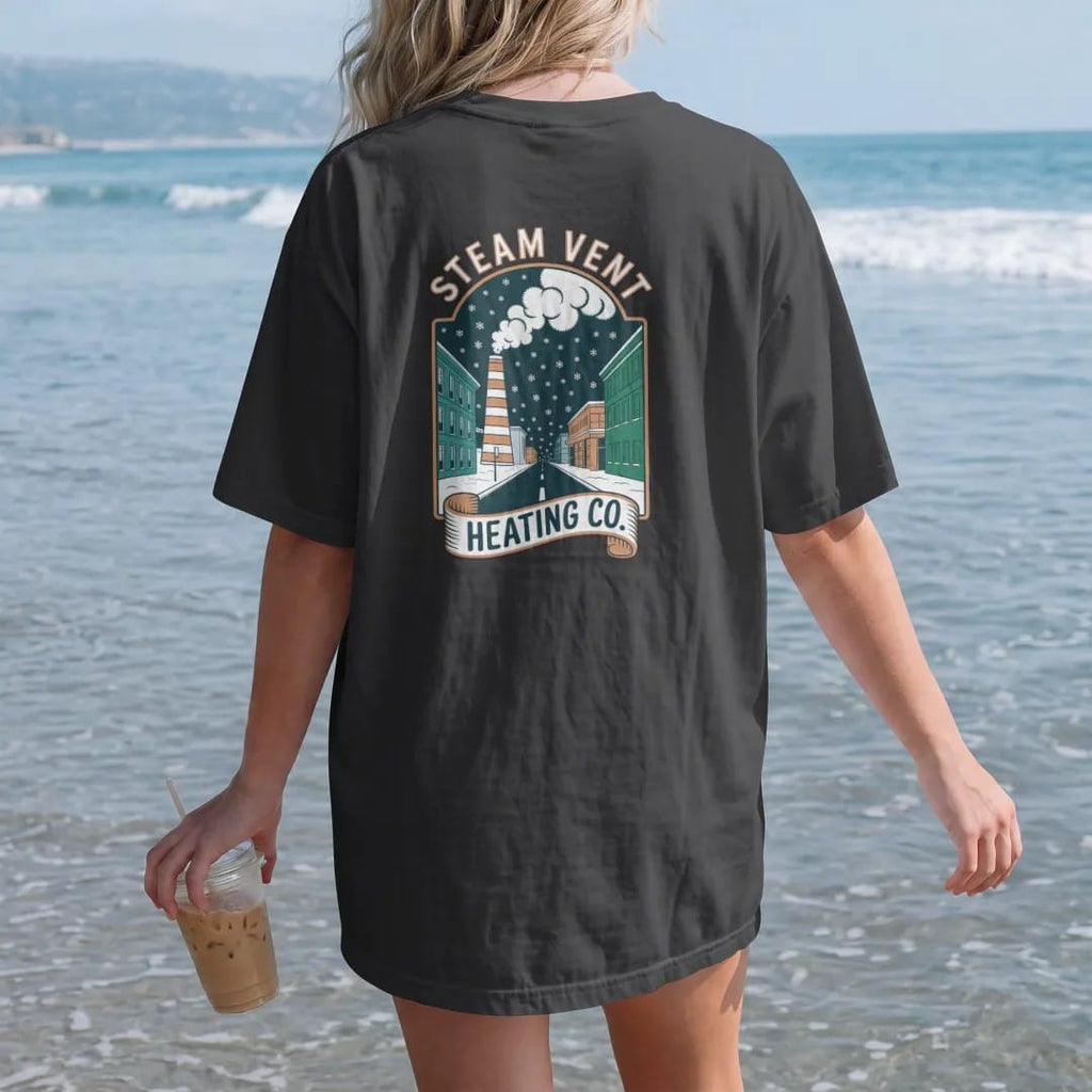 Woman on the beach in a Black tee, back view: a striped steam stack puffing clouds in falling snow with Steam Vent arched above a Heating Co. ribbon, in bone white, orange, yellow and green.