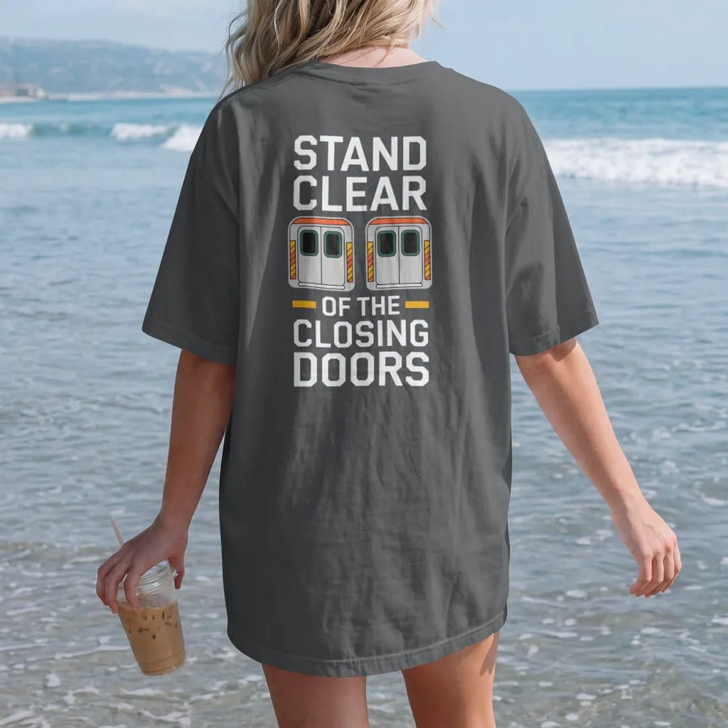 Woman on the beach in a Graphite tee, back view: two closing subway doors with a warning stripe under Stand Clear and Of The Closing Doors in transit-style lettering, in bone white, yellow, orange and green.