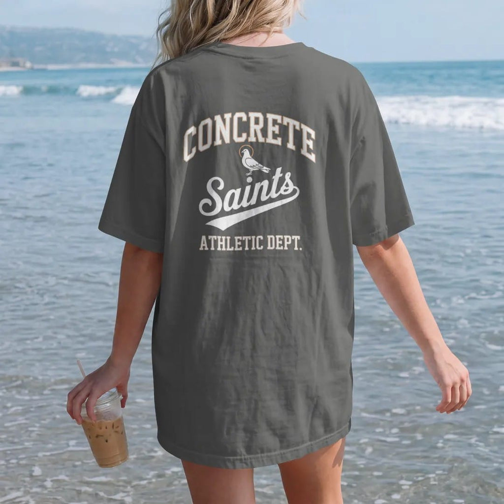 Woman on the beach in a Pepper tee, back view: a varsity layout with Concrete arched over a Saints script flourish, Athletic Dept. below and a haloed pigeon mascot, in bone white, orange and green.