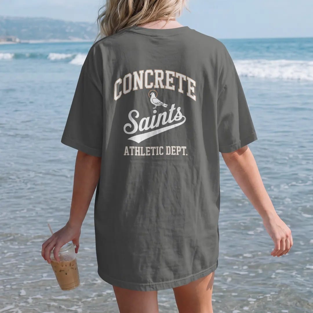 Woman on the beach in a Pepper tee, back view: a varsity layout with Concrete arched over a Saints script flourish, Athletic Dept. below and a haloed pigeon mascot, in bone white, orange and green.
