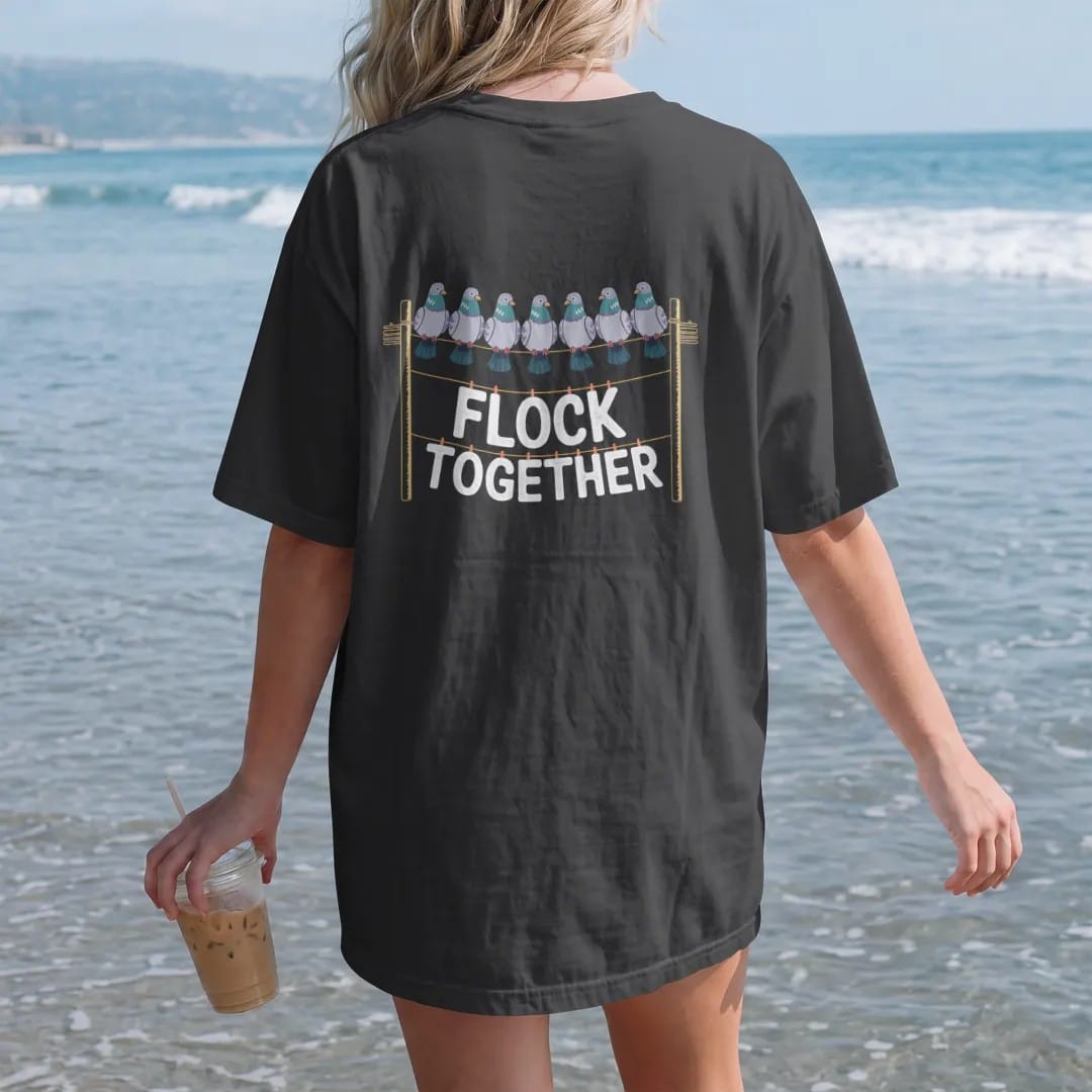 Woman on the beach in a Black tee, back view: a row of pigeons on a power line between two poles with Flock Together below, in bone white, green, orange and yellow.