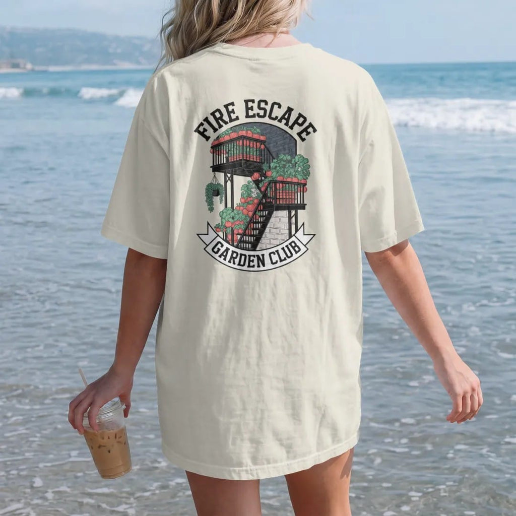 Woman on the beach in a Ivory tee, back view: a fire escape crowded with potted tomatoes and herbs under Fire Escape and over a Garden Club banner, in charcoal, brick red, green and navy.