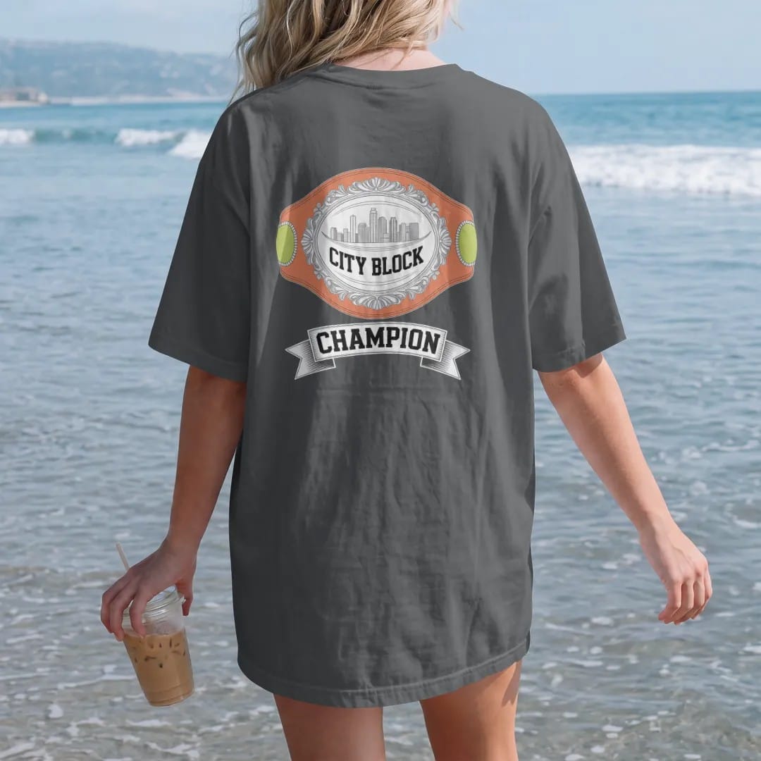 Woman on the beach in a Graphite tee, back view: a heavyweight championship belt with a skyline plate reading City Block over a Champion banner, in yellow, bone white, orange and green.