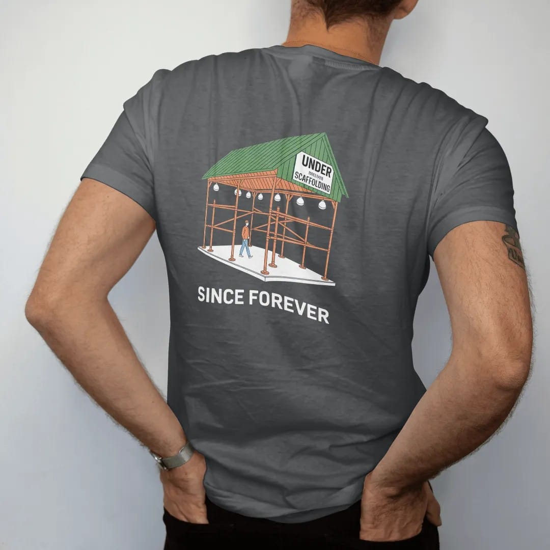 Man wearing a Graphite tee, seen from the back: a green sidewalk scaffolding shed with work lights over a tiny walker, labeled Under Scaffolding and Since Forever, in green, yellow, orange and bone white.