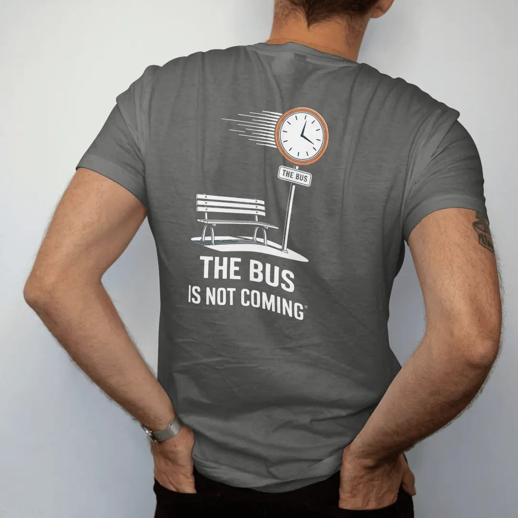 Man wearing a Pepper tee, seen from the back: a bus stop sign over an empty bench and spinning clock reading The Bus Is Not Coming, in bone white, orange, yellow and green.