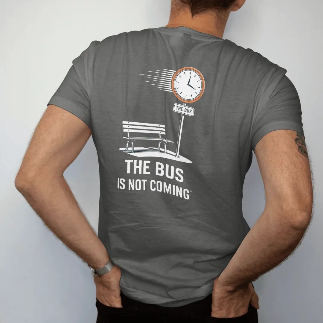 Man wearing a Pepper tee, seen from the back: a bus stop sign over an empty bench and spinning clock reading The Bus Is Not Coming, in bone white, orange, yellow and green.