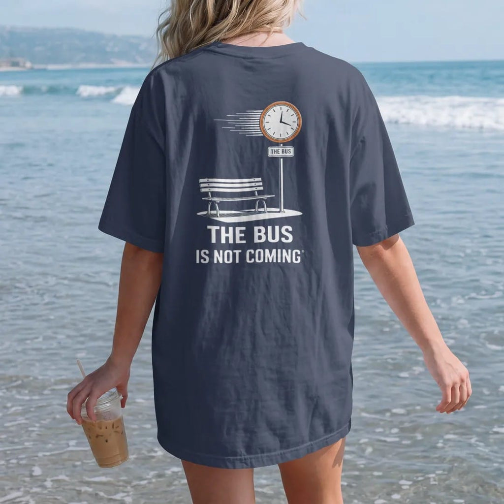Woman on the beach in a Navy tee, back view: a bus stop sign over an empty bench and spinning clock reading The Bus Is Not Coming, in bone white, orange, yellow and green.