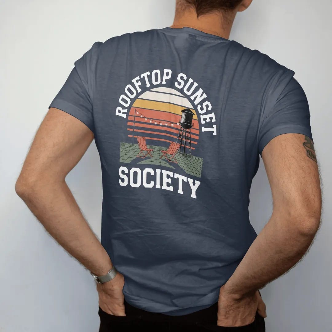 Man wearing a Midnight tee, seen from the back: two folding chairs and string lights on a rooftop facing a striped sunset and water tower, under Rooftop Sunset and over Society, in orange, yellow, bone white and green.