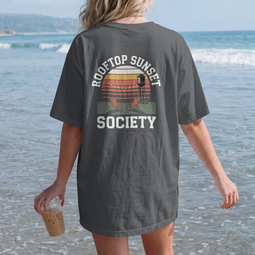 Woman on the beach in a Graphite tee, back view: two folding chairs and string lights on a rooftop facing a striped sunset and water tower, under Rooftop Sunset and over Society, in orange, yellow, bone white and green.