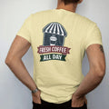 Man wearing a Butter tee, seen from the back: a steaming paper coffee cup in front of a striped awning with Fresh Coffee arched above an All Day ribbon, in charcoal, brick red, green and navy.
