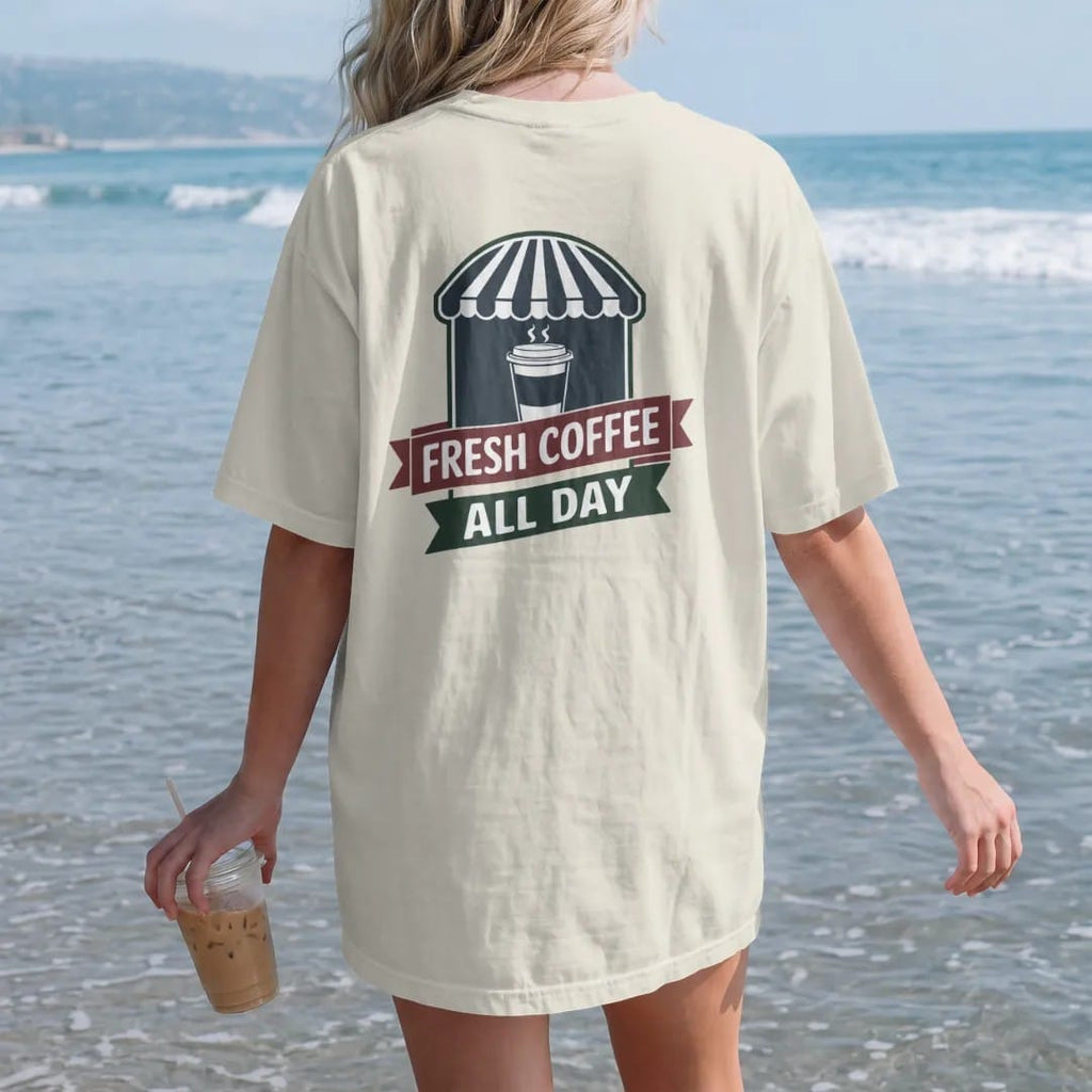 Woman on the beach in a Ivory tee, back view: a steaming paper coffee cup in front of a striped awning with Fresh Coffee arched above an All Day ribbon, in charcoal, brick red, green and navy.