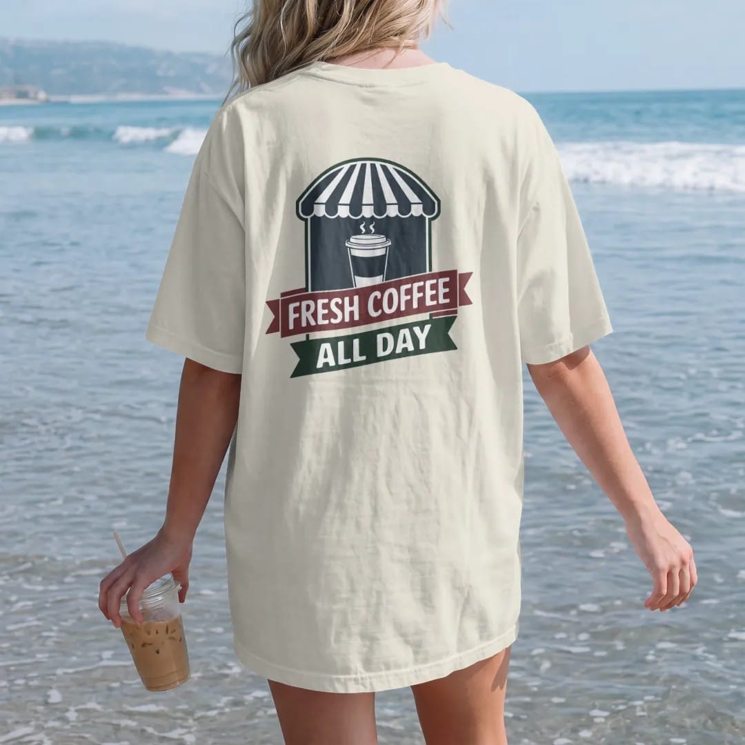 Woman on the beach in a Ivory tee, back view: a steaming paper coffee cup in front of a striped awning with Fresh Coffee arched above an All Day ribbon, in charcoal, brick red, green and navy.