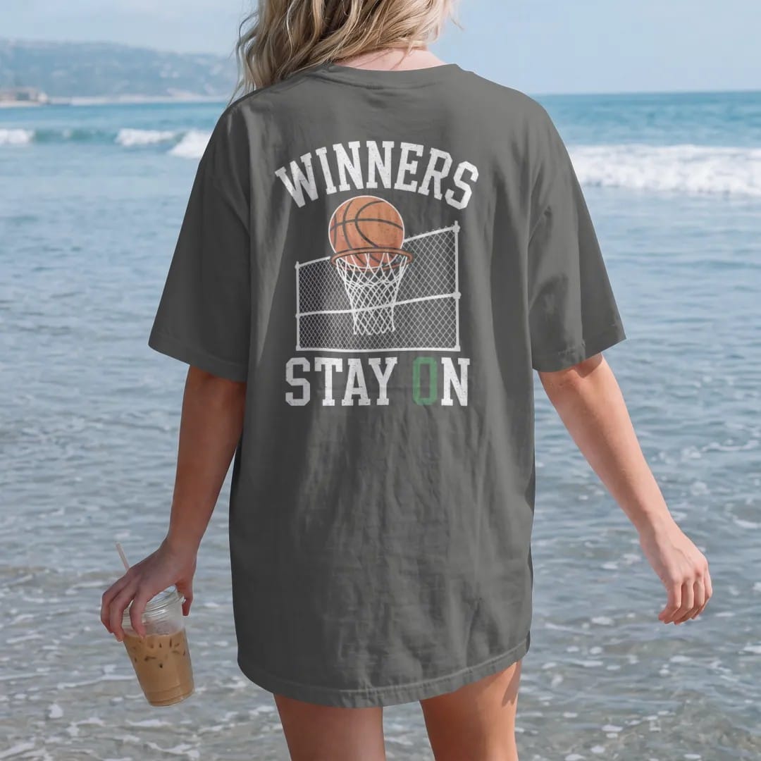 Woman on the beach in a Pepper tee, back view: a basketball swishing a chain net on a fenced street court under an arched Winners over Stay On, in orange, bone white, yellow and green.