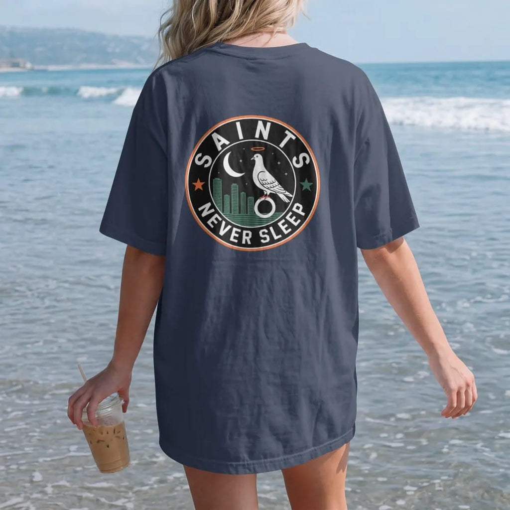 Woman on the beach in a Navy tee, back view: a round badge with a night skyline, crescent moon and haloed pigeon, ringed by Saints and Never Sleep, in bone white, yellow, orange and green.