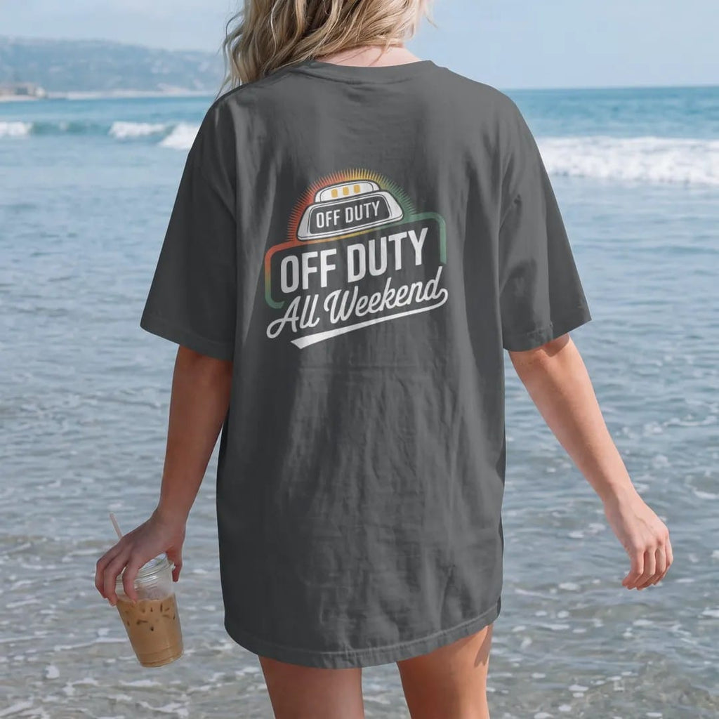 Woman on the beach in a Graphite tee, back view: a glowing taxi roof light reading Off Duty above an All Weekend script banner, in yellow, orange, bone white and green.