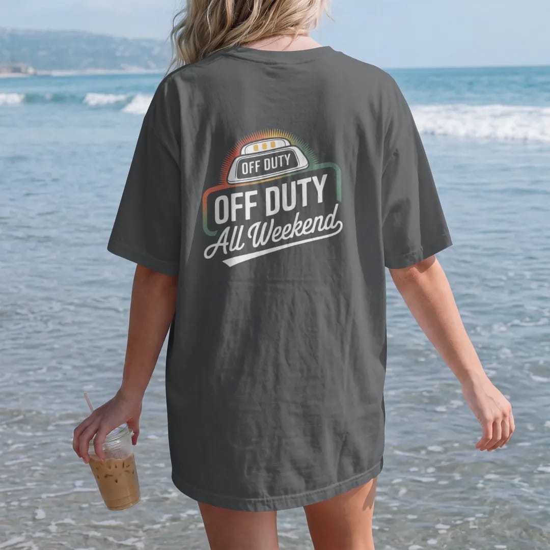 Woman on the beach in a Graphite tee, back view: a glowing taxi roof light reading Off Duty above an All Weekend script banner, in yellow, orange, bone white and green.