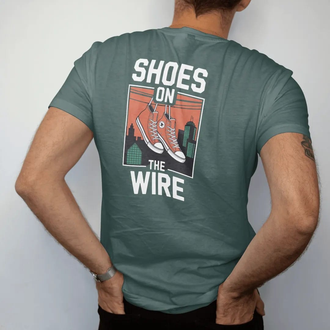 Man wearing a Blue Spruce tee, seen from the back: a pair of high-top sneakers hanging by laces from a power line over rooftops, with Shoes On above and The Wire below, in bone white, orange, green and yellow.