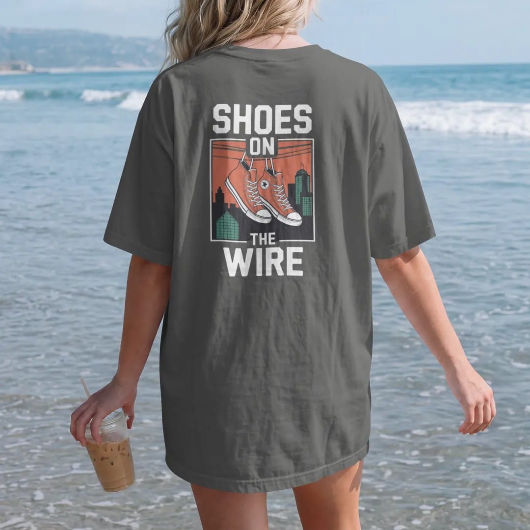 Woman on the beach in a Pepper tee, back view: a pair of high-top sneakers hanging by laces from a power line over rooftops, with Shoes On above and The Wire below, in bone white, orange, green and yellow.