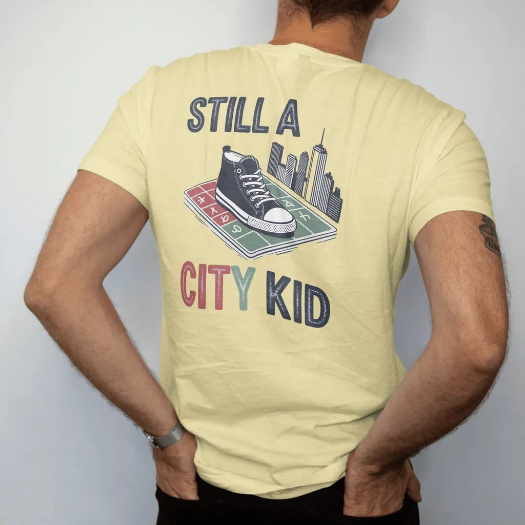 Man wearing a Butter tee, seen from the back: a sidewalk square with a sneaker print, chalk hopscotch and skyline doodle under Still A and City Kid in chalky lettering, in charcoal, brick red, green and navy.
