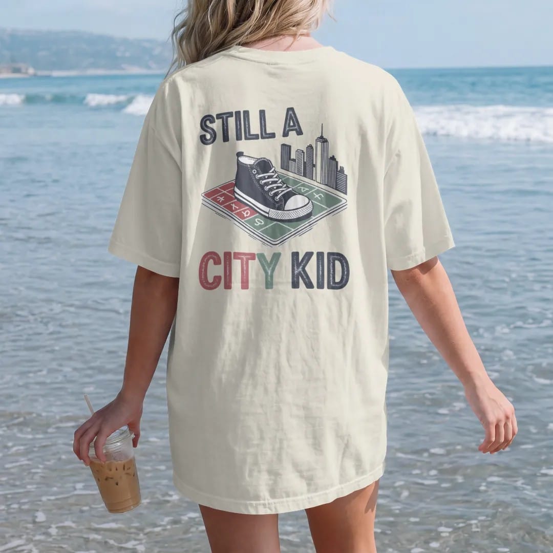 Woman on the beach in a Ivory tee, back view: a sidewalk square with a sneaker print, chalk hopscotch and skyline doodle under Still A and City Kid in chalky lettering, in charcoal, brick red, green and navy.