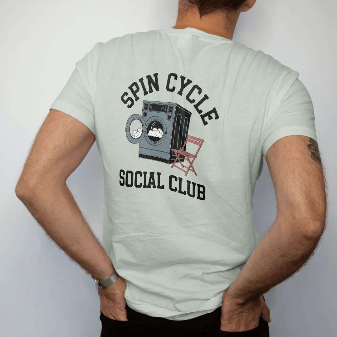 Man wearing a Bay tee, seen from the back: a retro laundromat washer with spinning suds and a folding chair under Spin Cycle and over Social Club, in charcoal, brick red, green and navy.