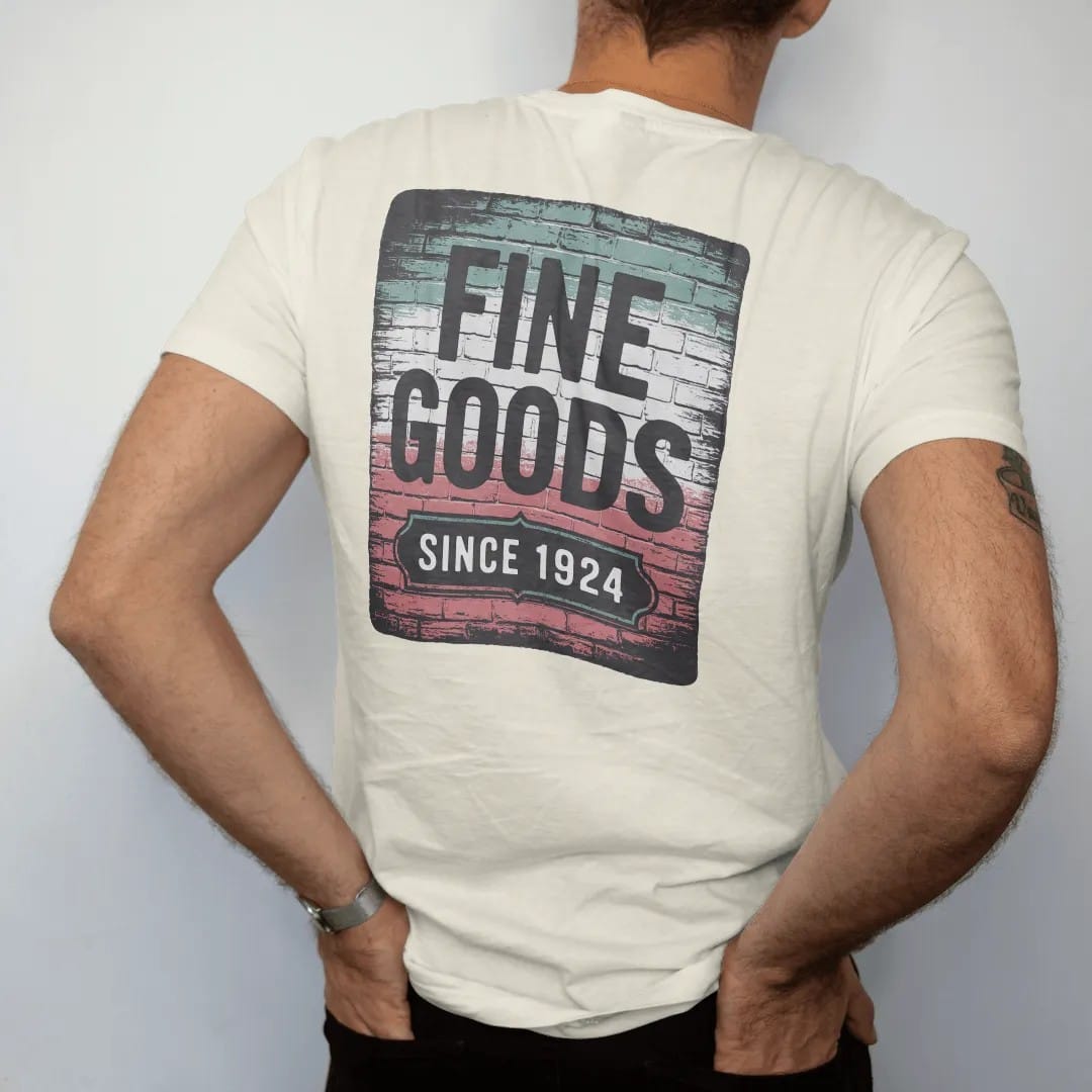Man wearing a Ivory tee, seen from the back: a faded painted brick-wall ad reading Fine Goods with a Since 1924 panel, brick texture showing through, in charcoal, brick red, green and navy.