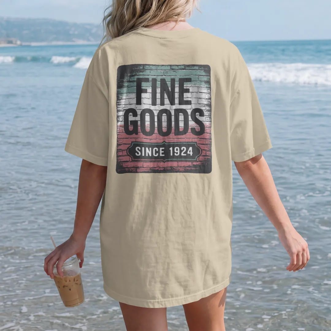 Woman on the beach in a Sandstone tee, back view: a faded painted brick-wall ad reading Fine Goods with a Since 1924 panel, brick texture showing through, in charcoal, brick red, green and navy.