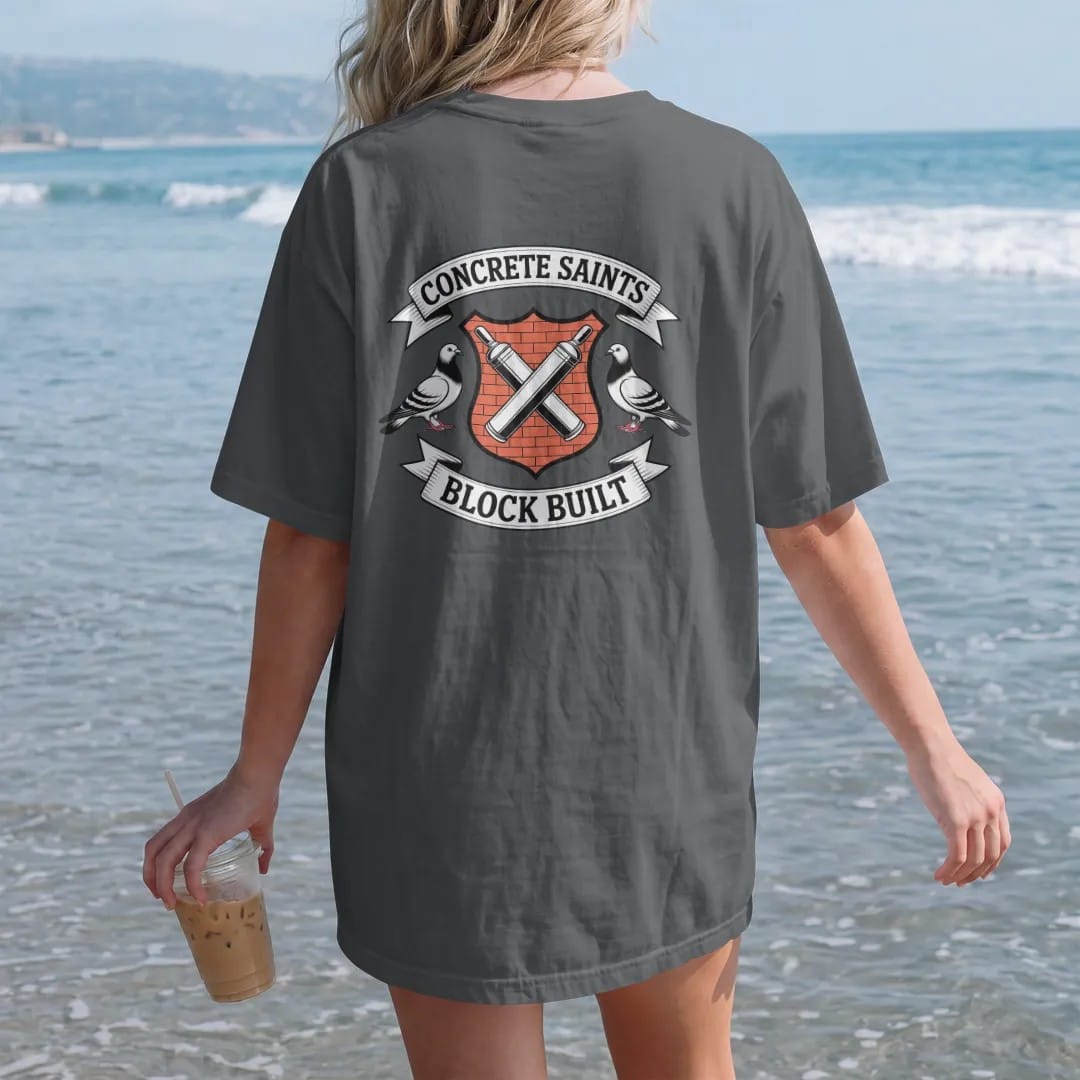 Woman on the beach in a Graphite tee, back view: a crest shield with crossed spray cans and two haloed pigeons, Concrete Saints on the top banner and Block Built on a ribbon, in bone white, orange, green and yellow.