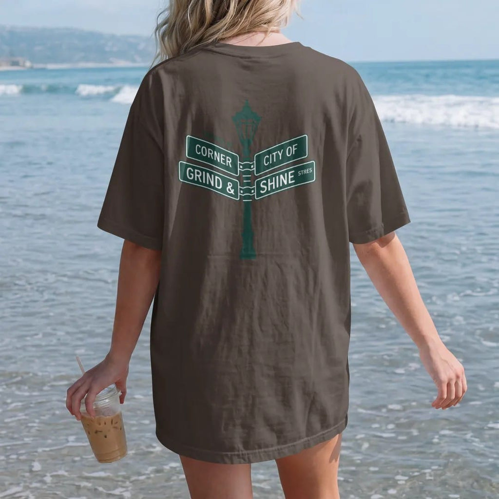 Woman on the beach in a Espresso tee, back view: two crossed street signs on a lamp post reading Grind and Shine under the words Corner Of, in green, bone white, yellow and orange.