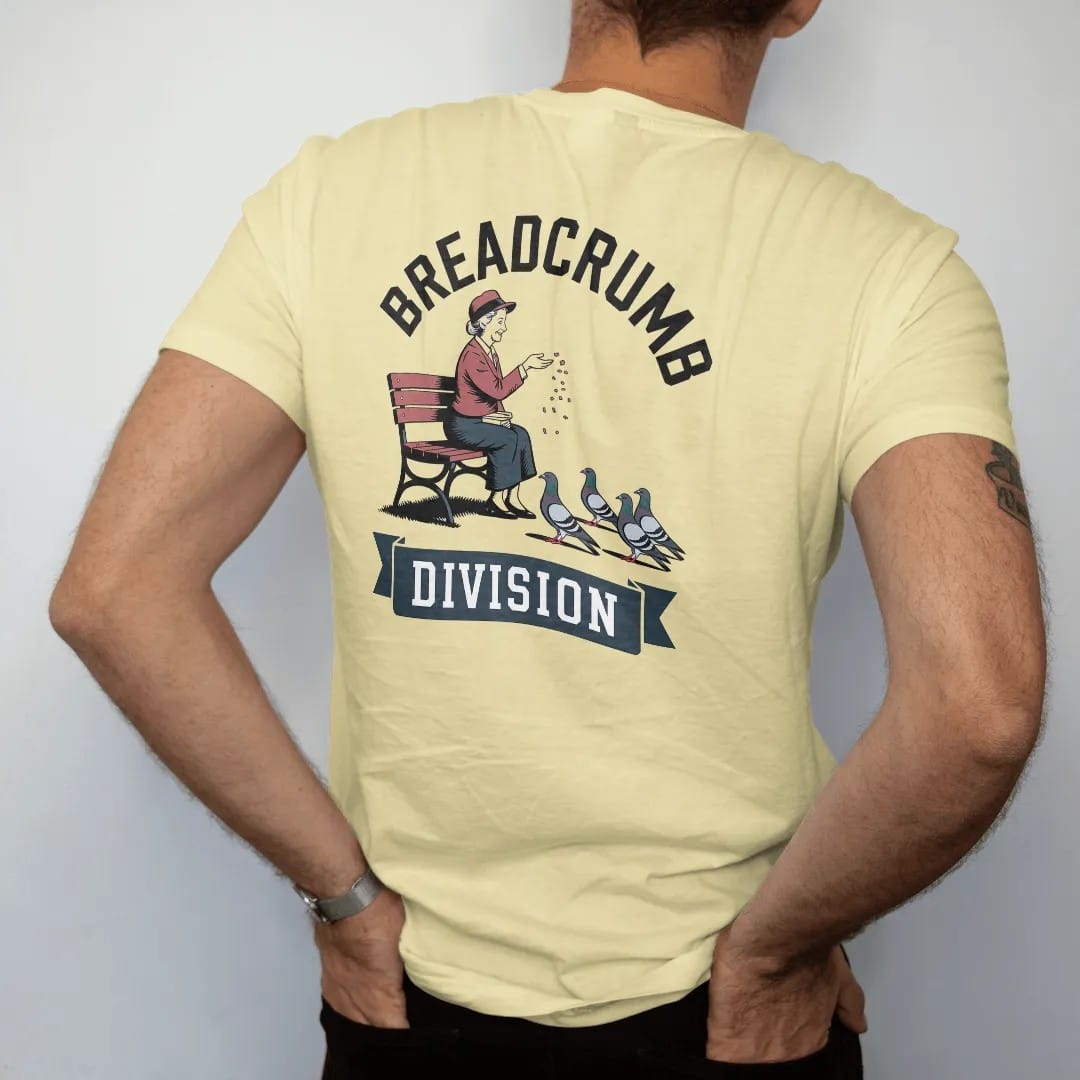 Man wearing a Butter tee, seen from the back: an older woman on a park bench tossing crumbs to pigeons under Breadcrumb arched over a Division ribbon, in charcoal, brick red, green and navy.