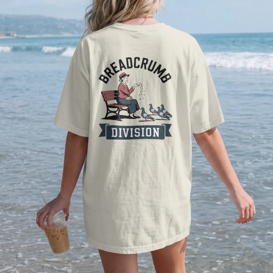 Woman on the beach in a Ivory tee, back view: an older woman on a park bench tossing crumbs to pigeons under Breadcrumb arched over a Division ribbon, in charcoal, brick red, green and navy.