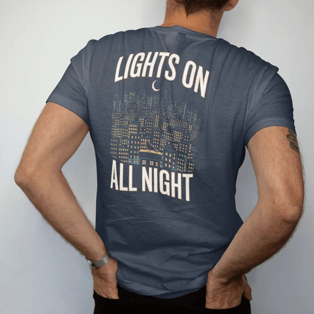 Man wearing a Midnight tee, seen from the back: a night skyline of lit windows under a crescent moon with a haloed pigeon on a ledge, framed by Lights On and All Night, in yellow, bone white, orange and green.