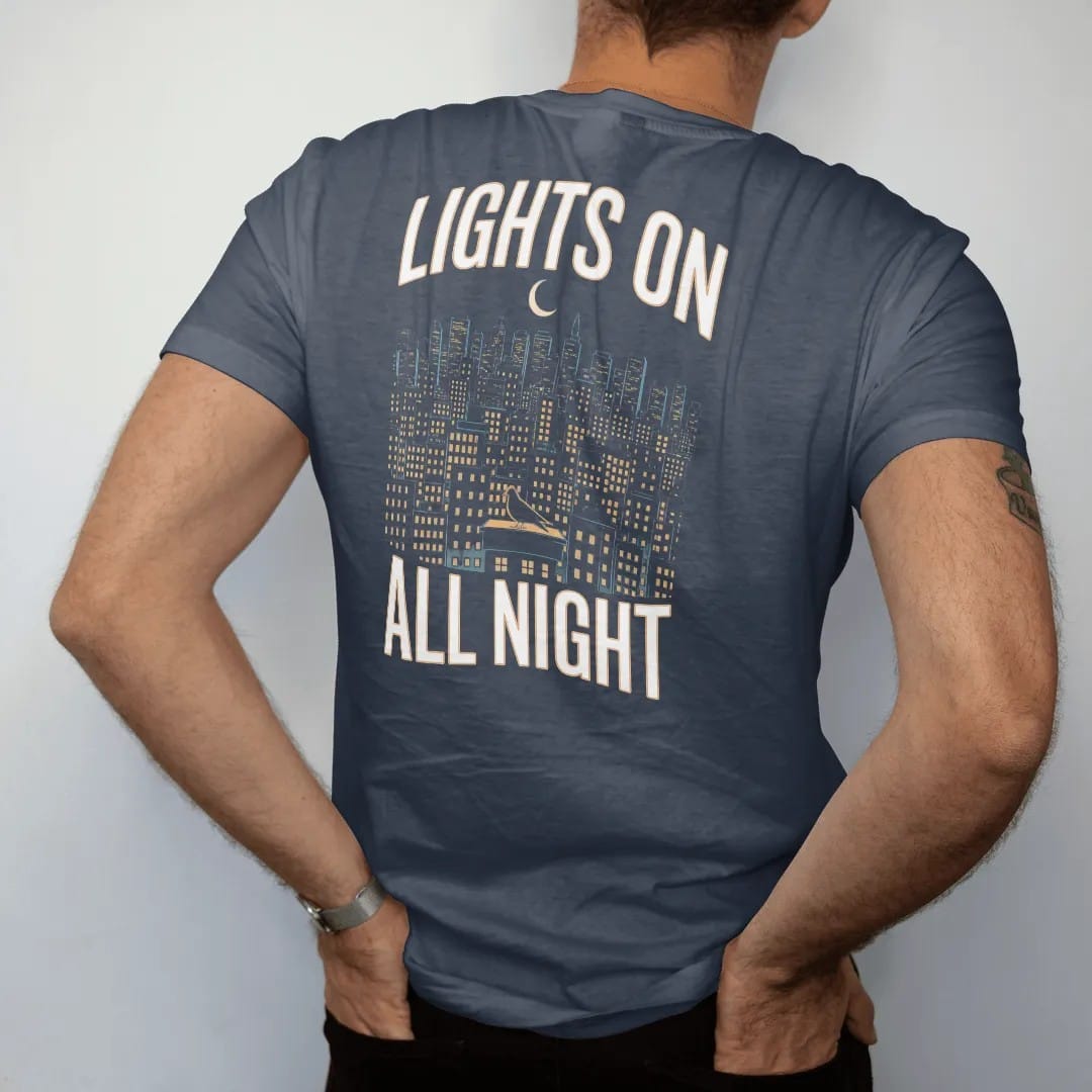 Man wearing a Midnight tee, seen from the back: a night skyline of lit windows under a crescent moon with a haloed pigeon on a ledge, framed by Lights On and All Night, in yellow, bone white, orange and green.