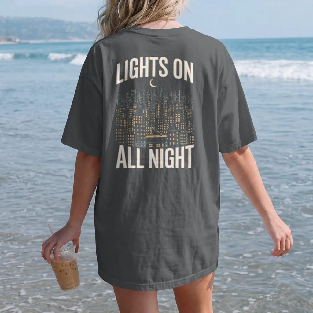 Woman on the beach in a Graphite tee, back view: a night skyline of lit windows under a crescent moon with a haloed pigeon on a ledge, framed by Lights On and All Night, in yellow, bone white, orange and green.