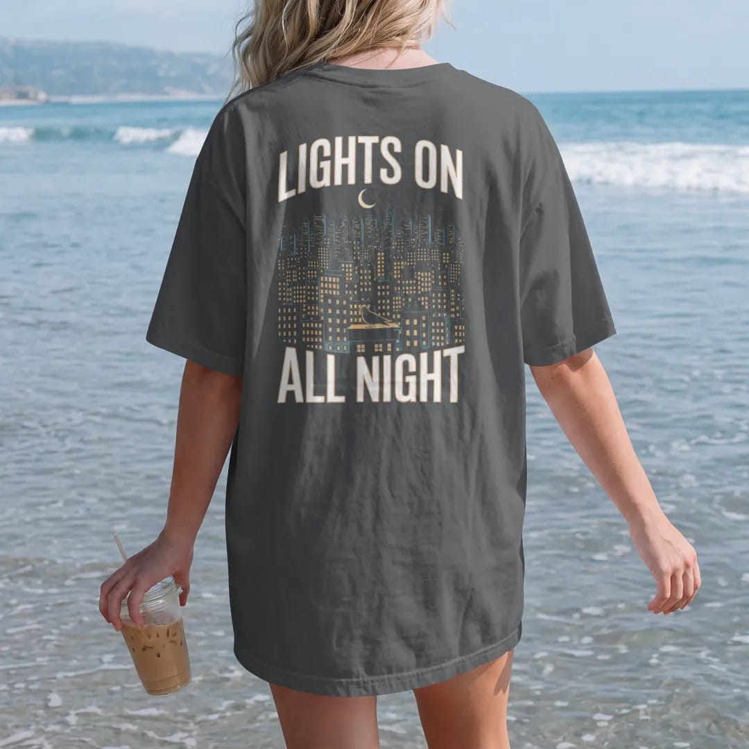 Woman on the beach in a Graphite tee, back view: a night skyline of lit windows under a crescent moon with a haloed pigeon on a ledge, framed by Lights On and All Night, in yellow, bone white, orange and green.